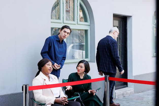 Crowd of diverse people sitting outdoors at shopping center door waiting in line to shop for seasonal holiday sales. Anxious customers at entrance of retail store wait for Black Friday shopping deals