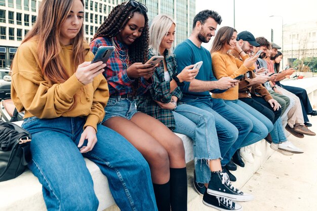 Толпа многонациональных молодых людей, сидящих в очереди на открытом воздухе, просматривающих киберпространство с мобильным телефоном