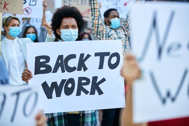 Crowd of people protesting against job loss due to COVID19 pandemic Focus is on African American woman holding a placard with back to work inscription