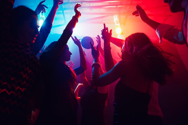 A crowd of people in silhouette raises their hands on dancefloor on neon light background. Night life, club, music, dance, motion, youth. Purple-pink colors and moving girls and boys.