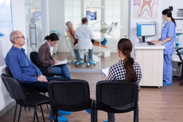 Crowded stomatology waiting area with people filling form for dental consultation. Stomatoloy specialist denstiry treating senior woman cavity. Receptionist working on computer.