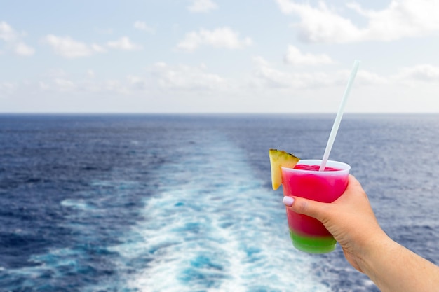 Cruise ship wake or trail on ocean surface with hand holding a glass with cocktail and straw