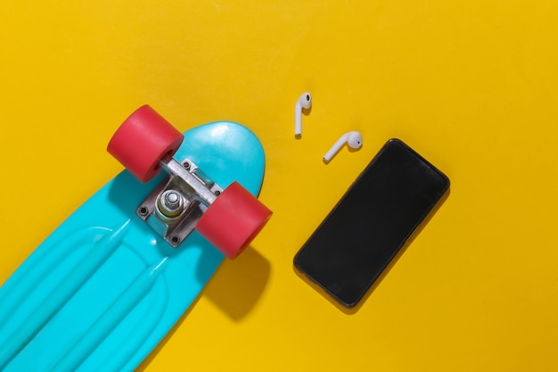 Cruiser board, smartphone and wireless headphones on a bright yellow background. Youth accessories, hipster outfit.