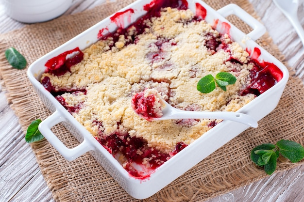 Crumble with strawberries in white dish on white wooden background