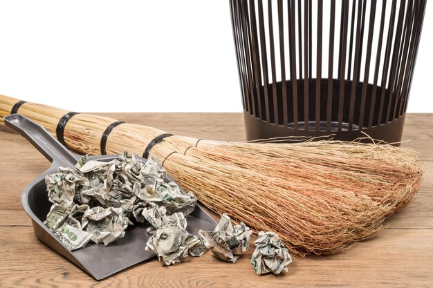 Crumpled dollars in a garbage shovel next to a broom and a bucket on a wooden surface on a white background