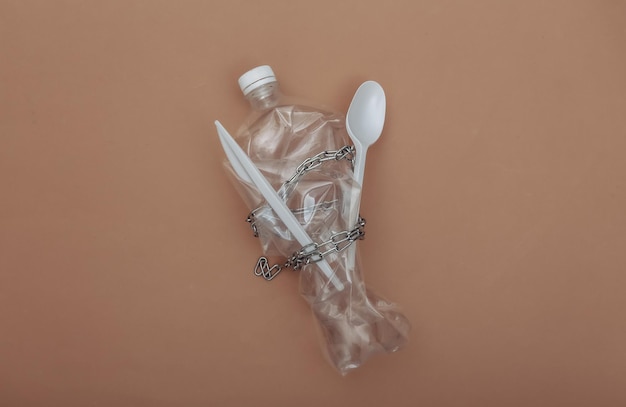 Crumpled plastic bottle, knife and spoon wrapped in a steel chain on brown background. Eco concept. Top view