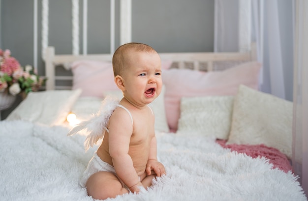 Crying baby in a diaper and angel wings sits on the bed in the room