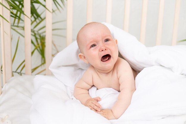 A crying baby girl in a blanket in a crib on a white cotton bed of six months has woken up and is calling for her mother