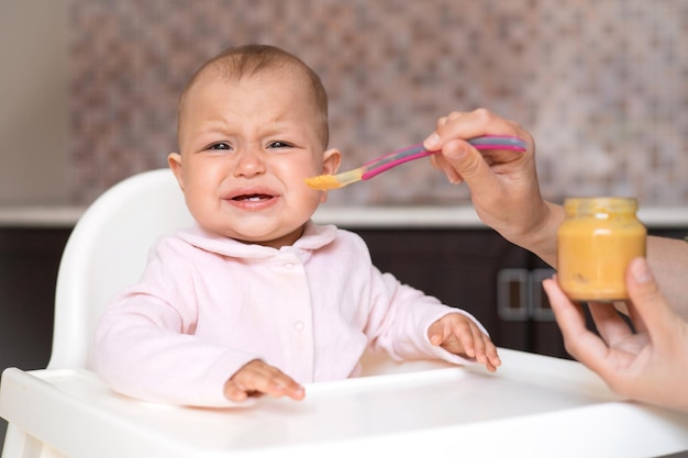 A crying baby refuses to eat baby food a cranky child real\
motherhood feeding the baby from a spoon vegetable and fruit puree\
kitchen