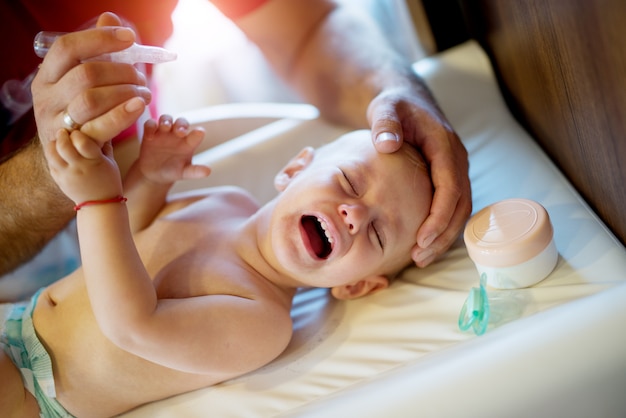 Crying beautiful baby kid lying on the bad with a pacifier and cream near the head and rejecting to get nose drops.
