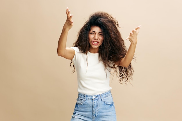 Crying beautiful latin female in basic white tshirt with afro\
hold hands up scream at camera scandal concept studio shoot over\
pastel beige background copy space free place for design ad