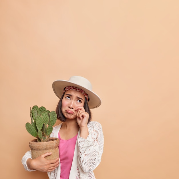 Crying doleful Asian woman rubs eyes wipes tears has frustrated expression focused above holds pot of cactus feels lonely and upset dressed in fashionable clothes isolated over beige wall