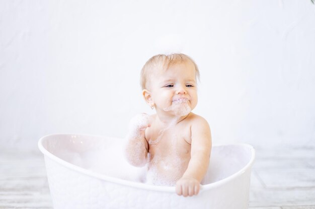 A crying little baby girl with foam in her mouth and in front of her eyes bathes in a bathtub with foam and soap bubbles in a bright bathroom, the concept of child hygiene