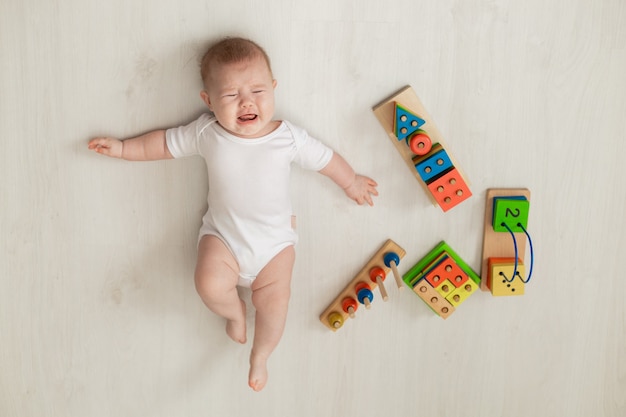 Crying newborn baby in white bodysuit lies on his back on the floor and plays with educational toys