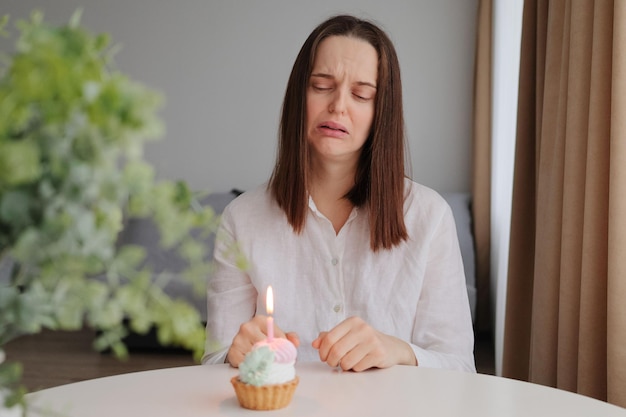 悲しい絶望を泣いている白人の茶色の髪の女性が家の内部で誕生日ケーキを持ってテーブルに座って、一人で祝うために動揺している不幸な表情でデザートを見ている