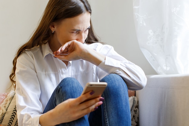 Piangere la donna in depressione, guardando la finestra, tenendo smartphone, coprendosi la bocca con la mano. relazione d'amore