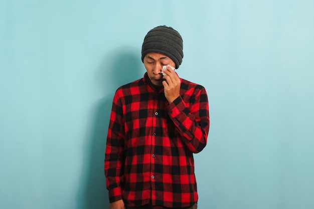 Photo crying young asian man wipes tears with a tissue while standing against a blue background