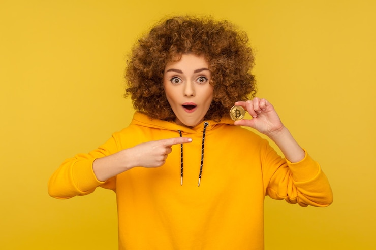 फोटो cryptocurrency, digital money. surprised excited curly-haired woman in urban style hoodie showing golden bitcoin and looking with amazement, pointing at btc coin. indoor studio shot, yellow background