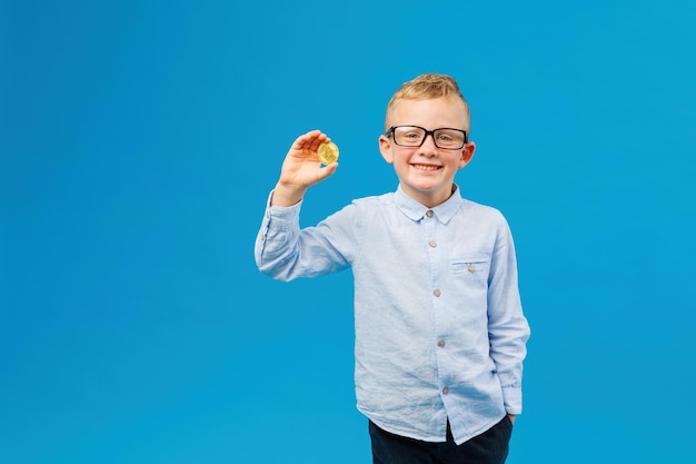 Cryptocurrency and modern finance Cheerful boy in eyeglasses holding golden bitcoin in studio on blue background