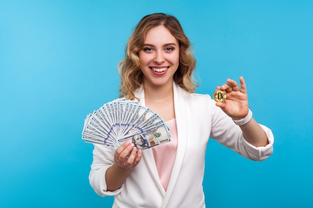 Cryptocurrency. Portrait of glad charming woman with wavy hair in white jacket holding physical golden bitcoin and dollars, advertising of btc crypto coin and cash. indoor studio shot, blue background