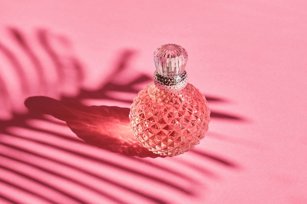 Bottiglia di profumo di cristallo con una bottiglia d'argento su sfondo rosa, ombra di una foglia di palma, acqua di toilette rosa, profumo femminile