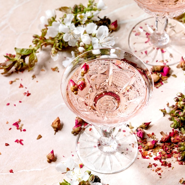 Crystal glasses of pink rose champagne, cider or lemonade with dry rose buds. Blossom cherry branches above. Pink marble background. Square image