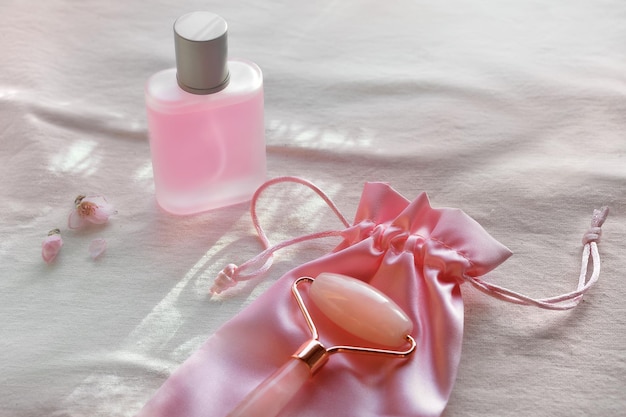 フェイスマッサージ用クリスタルローズクォーツフェイシャルローラーピンクのスキンケア製品またはすりガラスの瓶に入った香水オフホワイトのテキスタイル背景自然光