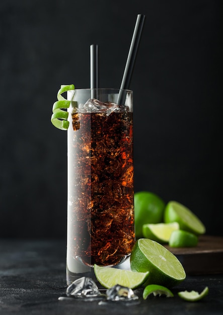 Cuba Libre cocktail in highball glass with ice and lime peel with straw and fresh limes on black table background