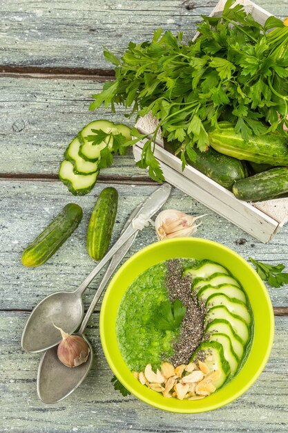 Cucumber Gazpacho. Green fresh cold summer soup. Fresh vegetables, greens, spices and cutlery. Old wooden boards background, top view