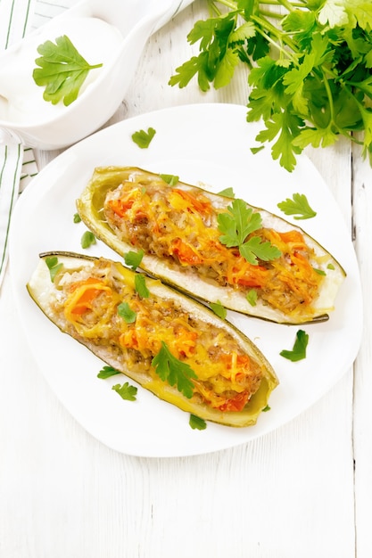 Cucumbers stuffed with minced meat, tomatoes, peppers with cheese and parsley in plate, cream and greens sauce in a gravy boat, a towel on wooden board background from above