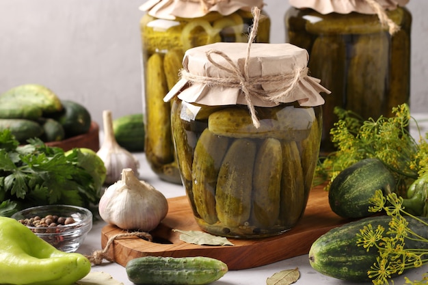 Cucumbers with garlic, pepper and dill in a glass jars on light grey background with veggies around