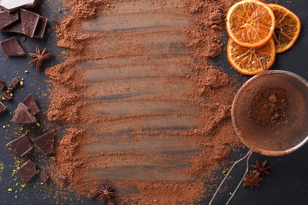 Culinaire zoete achtergrond. Gezeefd aromatisch cacaopoeder verspreid over zwarte leisteen met sinaasappelcitrusschijfjes en gebroken chocoladerepen. Ingrediënten voor het maken van brownies. Bovenaanzicht, kopieer ruimte