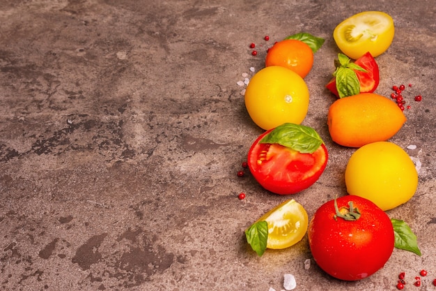 Culinary background with ripe tomatoes and basil leaves. Various colorful vegetables, sea salt, and pink peppercorn on a dark stone concrete background, copy space