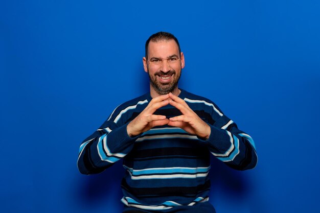 Cunning bearded man clasping hands and planning evil tricky prank or scheming cheating with sly smile indoor studio shot isolated on blue background