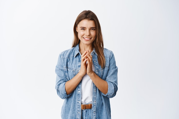 Astuta giovane donna bionda con le dita del campanile e sorridente, prepara un piano, progetta qualcosa con una faccia pensierosa, pensa e fissa davanti, muro bianco