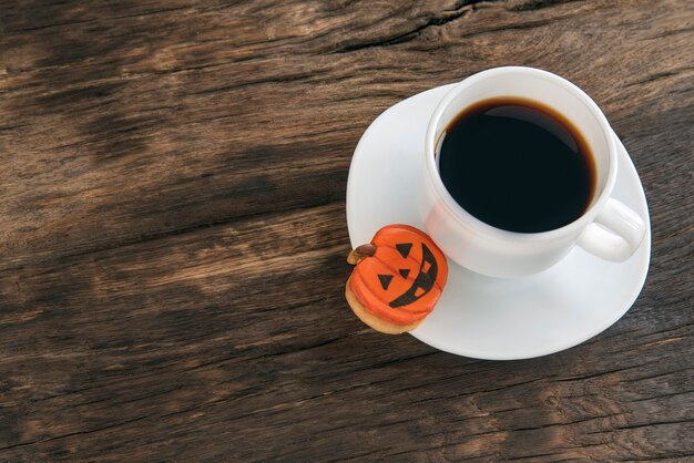 Cup of black coffee and a Halloween cookie-pumpkin stands on a brown wooden table. Halloween food concept. Copy space