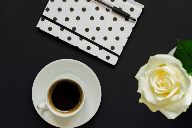 Cup of black coffee, notebook and white rose on