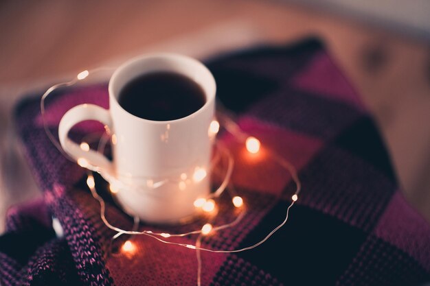 Cup of black tea staying on wool knitted sweaters over Christmas lights at background. Winter season.