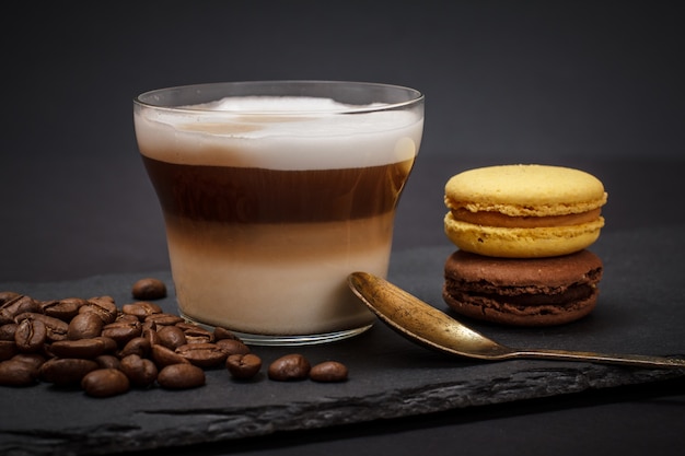 Cup of cappuccino, coffee beans, spoon and macaroons on black background.