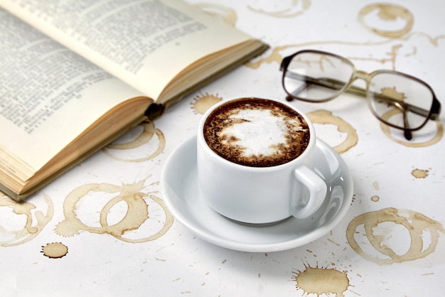 Cup of cappuccino coffee reading glasses and book on white table with traces of coffee cups