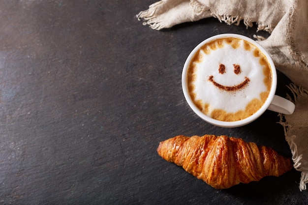 Cup of cappuccino coffee with drawing smile on milk foam and croissant