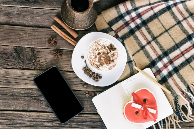 Tazza di cappuccino, messaggio di larghezza dei biscotti a forma di cuore, smartphone e caffettiere su una tavola di legno marrone