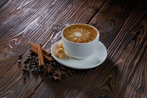 A cup of cappuccino with coffee beans, cinnamon and brown sugar on wooden table