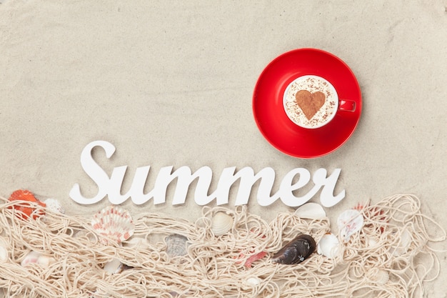 Cup of Cappuccino with heart shape symbol, word Summer and net with shells on sand