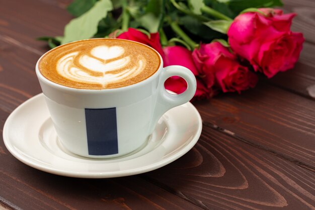 Cup of cappucino with art and bouquet of roses on wooden table