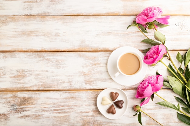 Cup of cioffee with chocolate candies, pink peony flowers on white wooden background. top view, 