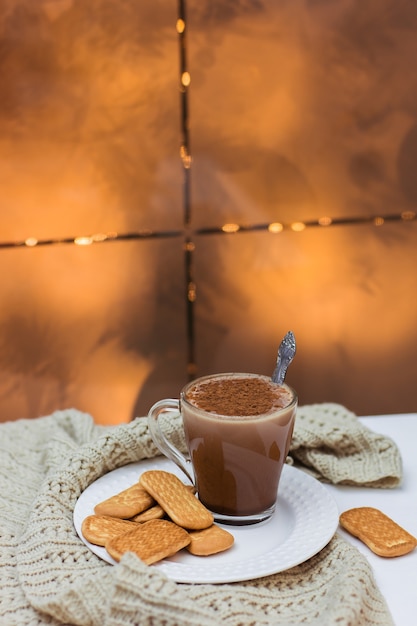 A cup of cocoa and cookies on a white plate on a white table and a knitted scarf