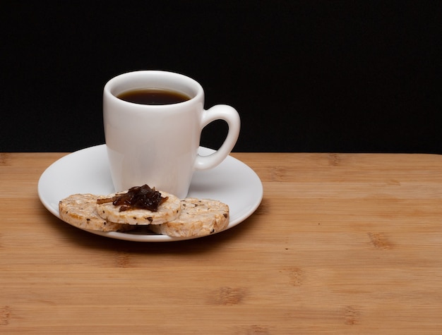 木のテーブルの下の上部にゼリーが付いているライスビーガンクッキーの横にあるコーヒーのカップ