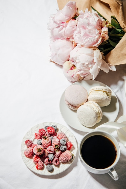 a cup of coffee berries macaroons and peony flowers on the bed