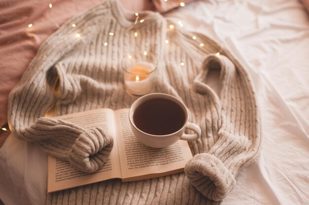 Cup of coffee or black tea staying on open book with burning candle and knitted gray sweater in bed